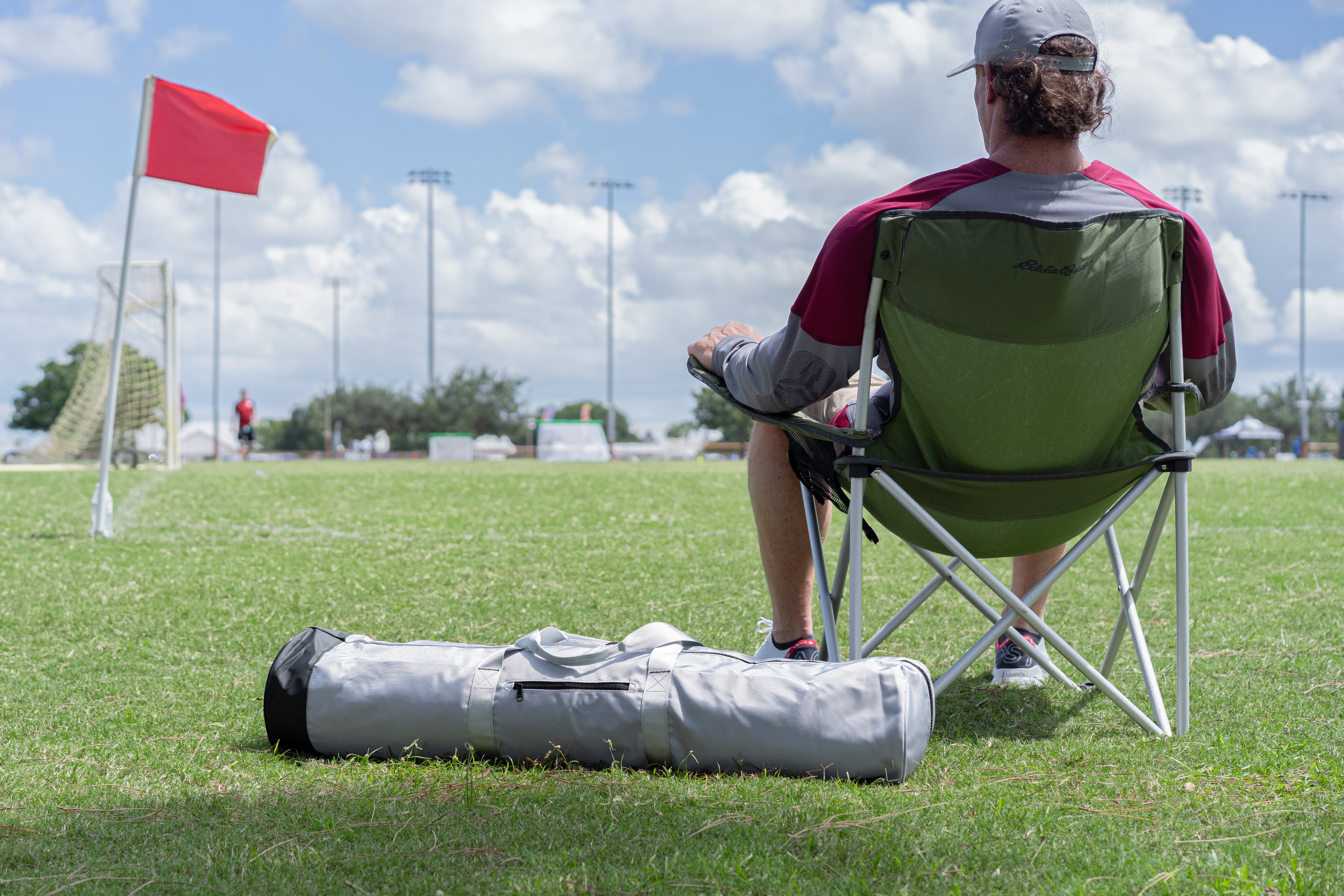 Person sitting on a green chair with a fieldlux bag by a soccer field.